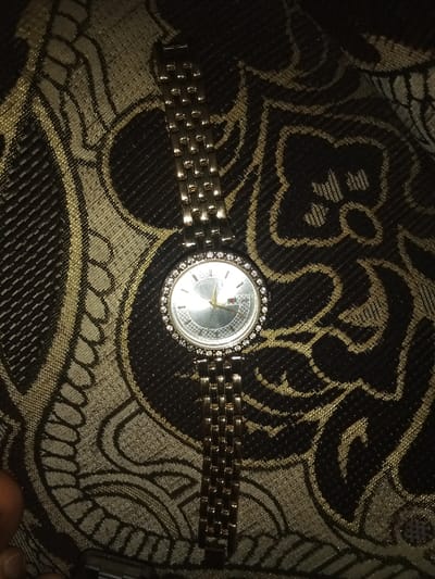 The image displays a gold-toned wristwatch positioned on a patterned textile surface. The watch features a circular silver-toned dial, enclosed by a bezel encrusted with clear gemstones. The dial contains three gold-toned hands and a visible brand emblem, consisting of the word "GUCCI" and a small rectangular motif with red, white, and blue segments. The watch's metallic link bracelet extends upwards and downwards from the watch face. The background fabric is dark, likely black or deep brown, adorned with a repeating pattern of intricate gold or beige curvilinear designs. The scene is dimly lit, resulting in a distinct glare on the watch's dial. A partial human finger, specifically a thumb, is visible in the bottom-left corner of the frame, alongside a small metallic object, indicating recent interaction with the watch. This scene is located in Umsobomvu Local Municipality, South Africa.