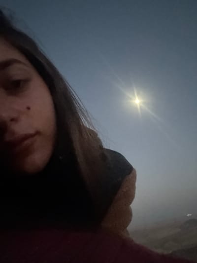 The original image, captured in Mitzpe Ramon, Israel, presents a partial, low-angle view of a person gazing up at a bright moon in a clear night sky. In the foreground, the lower left section features part of a person's face, specifically the cheek, nose, and mouth area, with dark hair visible along the top of the head. A distinct small mole is visible on the cheek. The person appears to be wearing warm attire, possibly a dark garment and a light brown, textured blanket or hood, which covers the right side of their head and shoulder.
The upper and central parts of the frame are dominated by a dark blue-grey sky, indicative of nighttime or very early morning. A brilliant, full moon shines prominently, displaying a clear starburst effect, which suggests a cloudless sky. The high contrast between the luminous moon and the dimly lit foreground creates a dramatic atmosphere. The scene evokes a moment of celestial observation, possibly a selfie or a casual photograph taken while admiring the night sky. Given the location context of Mitzpe Ramon, known for its desert environment and exceptional stargazing conditions, this image perfectly encapsulates the experience of witnessing the moon's beauty. No text is visible in the media file.