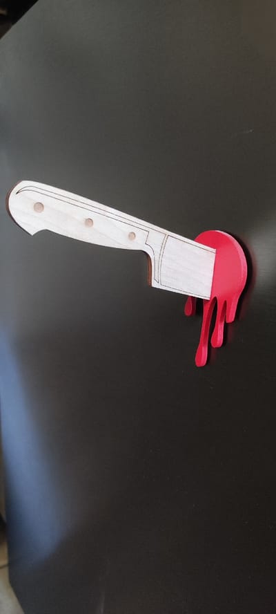 The image shows a 3D printed object shaped like a knife with a wooden handle and a red, dripping blade, attached to a dark, textured surface. The object is positioned horizontally. There is no discernible location in the city of Dinskaya, Russia, nor any people, actions, or interactions depicted. The environment appears to be a plain, dark surface.