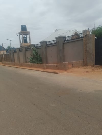 In Gindi Nassarawa Kyarmi Galbi, Nigeria, an image captures a segment of a street adjacent to a walled property. The wall, constructed from concrete blocks and rendered with cement, features decorative metalwork along its upper edge. A tall, concrete water tower stands behind the wall, supporting two black water tanks. A solar-powered street light is visible to the left of the water tower. The foreground shows an asphalt road, with a sandy verge running alongside the wall. A sparse, leafy tree grows within the property. The sky is overcast.