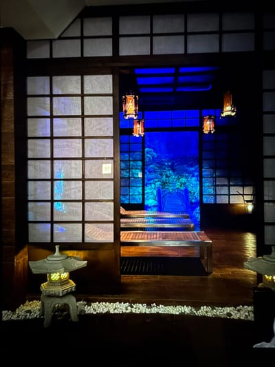 This image depicts an indoor scene that appears to be a themed exhibit or a dimly lit, decorative space. The architecture suggests a Japanese influence, with shoji-style screens framing a view into another area. In the foreground, to the left, is a stone lantern with a glowing light inside, placed on a bed of white pebbles. To the right, a wooden bench with metal trim is visible, extending towards the back of the scene.
The background features a vibrant blue illuminated mural or projection, depicting a lush, fantastical landscape with trees and a wooden bridge. Several paper lanterns with warm orange light hang from the ceiling, casting a soft glow. The floor is dark wood, reflecting the light from the lanterns and the mural.
There are no people or animals visible in the image. The overall impression is one of tranquility and artistic design. The location is unknown and there is no indication it is in Denver, Colorado, United States.