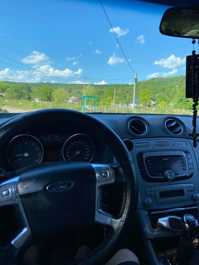 This is a photograph taken from inside a car, looking forward through the windshield. The car's dashboard, steering wheel with a Ford logo, and gear shift are visible in the foreground. The exterior scene shows a bright blue sky with scattered white clouds, rolling green hills covered in trees, and some houses. Power lines and a utility pole are also visible. The photograph appears to be taken in Negrești, Moldova, given the visual cues of a rural European landscape and the style of the buildings and infrastructure. The overall impression is one of a scenic drive on a clear day.