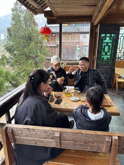 The scene depicts four individuals seated around a rustic wooden table on a covered veranda overlooking a traditional town in Gongtan, China. The veranda features exposed wooden beams, a wooden railing, and a red decorative lantern hanging from the ceiling. In the background, green trees and multi-story traditional wooden buildings with intricate latticework windows and dark roofs are visible under an overcast sky.

At the table, from left to right:
1.  A female, viewed from the back, has dark hair tied in a ponytail and wears a black jacket. Her right hand is near her mouth.
2.  A female, viewed in profile, wears a light-colored baseball cap with a "NY" logo and a dark jacket. She holds a light-colored smartphone in both hands, looking down at the screen. White decorative branches are positioned in front of her.
3.  A male, viewed in profile, wears a black leather jacket over a dark shirt. He gestures with his right hand near his mouth, appearing to speak. A black shoulder bag hangs on his side.
4.  A child, viewed from the back, has dark hair in a ponytail and wears a dark, puffy jacket with a visible grey hood underneath. The child is looking down at the table.

The wooden table holds a clear glass teapot containing green tea, several small teacups (some with tea, some empty), a disposable coffee cup with a lid, and multiple small plates/bowls filled with various snacks, including seeds and nuts. Two dark-colored smartphones and several smooth, dark stones are also on the table. The individuals appear engaged in a social gathering, enjoying tea and snacks in a relaxed, semi-outdoor setting.