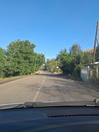 The image depicts a scene from a vehicle traveling along a road in Vayk, Armenia. The perspective is from inside the vehicle, looking out. The road is paved with a white line marking the center. On the left side of the road, there is a clear blue sky and a lush green tree. Further in the background, a hill is visible, and a wooden utility pole with wires stretches across the sky.

On the right side of the road, there are more trees and vegetation. A white fence with decorative metalwork is visible behind some of the trees, suggesting a residential property. The overall setting appears to be a rural or semi-rural area. The sunlight casts shadows on the road, indicating it is daytime. The weather appears to be clear and sunny. There is no discernible activity or event taking place, other than the implied movement of the vehicle. No people are visible in the image. No text is legible.