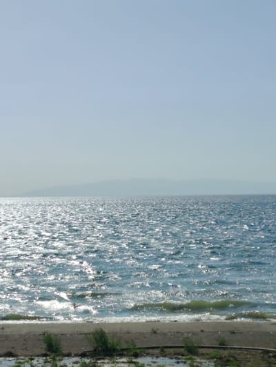 The image shows the coast of Lake Iznik in Iznik, Turkey. The water is choppy and reflecting sunlight, with small waves breaking on the shore. In the distance are mountains that form the lake's border. The beach consists of dark sand and gravel with some vegetation. The sky is clear and pale blue.