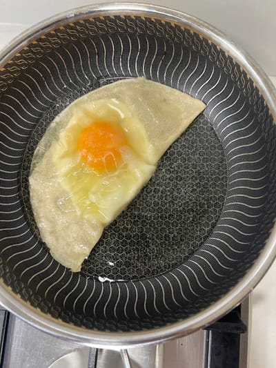 A crepe with an egg cooking in the center sits in a non-stick frying pan. The pan has a distinctive concentric, wavy pattern on its exterior. The crepe is folded in half, and the egg's yolk is visible in the center, surrounded by cooked egg white. The pan is on a stovetop, and there appears to be some oil or grease in the pan.