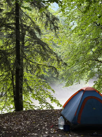 A blue and orange tent is pitched on a forest floor covered in leaves, next to a large body of water, possibly a river. A large plastic water bottle and a small blue container sit in front of the tent. The scene is surrounded by dense green foliage, with sunlight filtering through the leaves. The location is identified as Rozhkao, Russia.