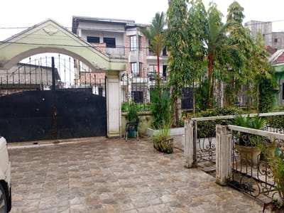 The image captures a view of a residential property entrance in Douala V, Cameroon. A large, dark metal gate with decorative scrollwork is partially open, revealing a glimpse of a paved courtyard. Above the gate, an arched structure painted in a light green hue with decorative plasterwork adorns the entrance. To the right of the gate, a white metal fence with ornate scrollwork encloses a garden area with various potted plants and lush greenery. Behind the fence, a multi-story building with light-colored walls and dark-rimmed windows is visible. In the foreground, the ground is covered with large, irregularly shaped stone-patterned tiles. A single black plastic chair sits to the left of the fence, near a large potted plant. Trees with dense green foliage are positioned strategically in front of the building and fence, contributing to a tropical atmosphere. The sky is overcast, suggesting a cloudy day.