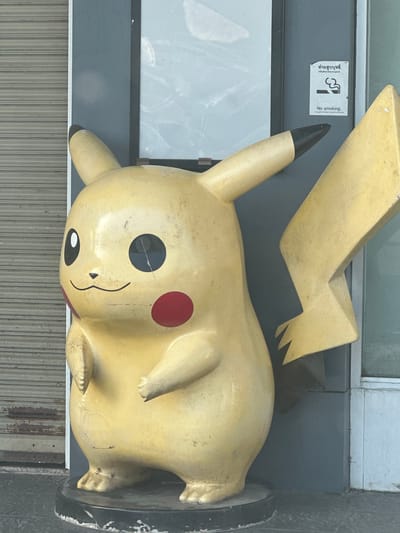 A weathered, large yellow Pikachu statue is positioned in front of a closed metallic storefront with a rolling shutter, located on Ban Ko Pru Road in Mueang Phuket, Thailand. The statue, which appears to be made of concrete or fiberglass, has faded paint and visible signs of wear, including chips and scuffs. One of Pikachu's large, black, circular eyes is cracked. To the right of the statue, a building facade features a gray panel with a "No Smoking" sign in both Thai and English, indicating a prohibition against smoking in the area. The ground in front of the statue is paved with dark tiles. The overall setting suggests an outdoor commercial area.