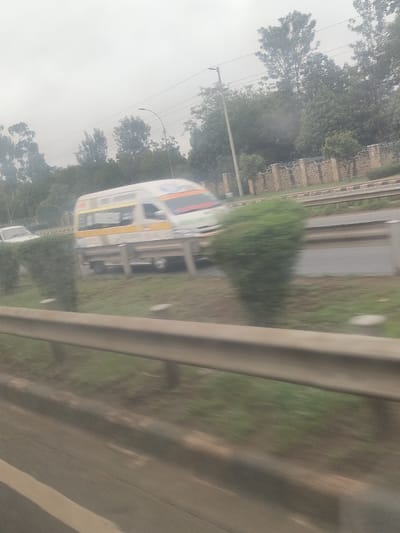 The image depicts a daytime scene in Gatongora ward, Kenya, captured from a moving vehicle, resulting in significant motion blur. In the foreground, a two-tier metallic guardrail runs horizontally. Beyond it, a grassy median with several trimmed, conical green bushes separates the lanes of a multi-lane paved road. A white minivan or minibus, adorned with yellow and orange horizontal stripes and an illegible sign on its windshield, is in motion on the road, traveling from right to left. Another white vehicle is partially visible further back on the left side of the road. In the background, a light-colored stone wall with dark decorative elements stretches horizontally, backed by dense green trees of varying heights. Utility poles with overhead lines and a streetlamp are interspersed among the trees. The sky is uniformly overcast and light grey. No individuals are distinctly visible.