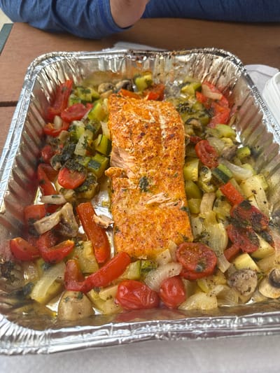 A cooked salmon fillet rests in a disposable aluminum foil pan filled with roasted vegetables. The salmon is seasoned with a reddish-orange spice blend and herbs, and is flaky and moist. The vegetables include cherry tomatoes, sliced zucchini, chunks of white onion, and mushrooms, all tossed with herbs and oil, and appear to have been roasted until tender and slightly caramelized. The pan is situated on a wooden surface, possibly a picnic table, and a person's arm, wearing a blue shirt, is visible in the upper left corner of the frame. The background is blurred, but suggests an outdoor setting. The image captures a meal ready to be served or consumed.