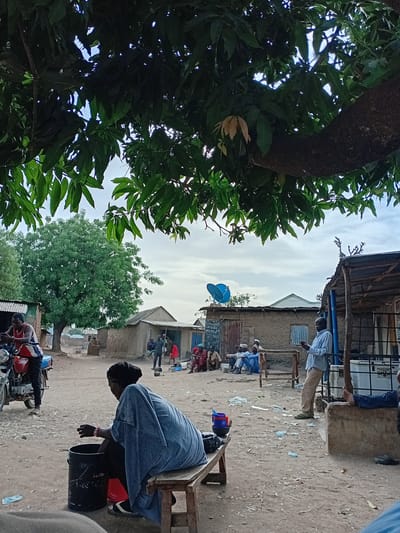 The image shows a street scene in Nata-Ala, Nigeria. A large tree with green leaves is on the left side of the frame. Several people are present. Some are seated, and one is leaning over a bucket while sitting on a wooden stool. A motorcycle is parked nearby. Buildings line the street, with a blue satellite dish visible on one. Various objects such as plastic bottles and containers are scattered on the ground.