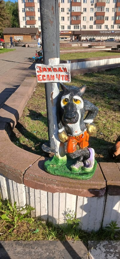 This media file shows a playful garden gnome in the shape of a wolf, lying on its back next to a pole. The gnome is black and white with large yellow eyes and is wearing an orange outfit and gold boots. It is positioned on a patch of grass with a red-brown concrete curb nearby. A white sign with red lettering is attached to the pole, reading "ЗАХОДИ ЕСЛИ ЧТО!" which translates to "COME IN IF YOU WANT!". In the background, a modern apartment building with multiple balconies can be seen, suggesting an urban or suburban setting. The scene appears to be outdoors on a sunny day, as indicated by the bright lighting and shadows. The overall impression is one of a whimsical and somewhat quirky outdoor decoration.