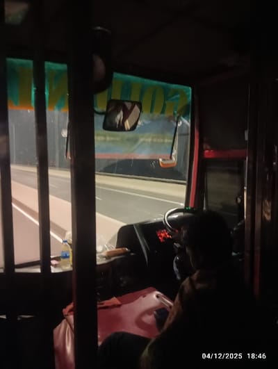 The image captures the interior of a large vehicle, likely a bus, traveling through the brightly lit Bangabandhu Sheikh Mujibur Rahman Tunnel in Naodoba, Bangladesh, at 18:46 on December 4, 2025. A solitary individual, presumably the driver, is seated in the foreground with their back to the camera, wearing dark attire and holding a mobile device in their right hand. The vehicle's dashboard displays an illuminated red digital readout. To the driver's right, a blue water bottle and packaged food items are visible on a small surface. Through the windshield, the tunnel's concrete structure and overhead green signage with yellow text, partially reflected, are clearly visible.