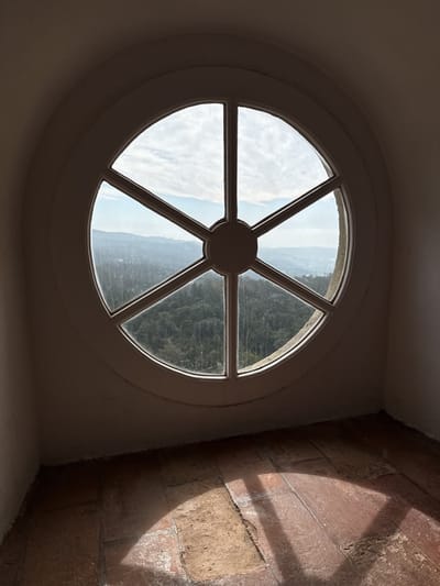 An eye-level, indoor shot depicts a six-paned circular window with a white frame situated within an arched opening. Outside the window, a landscape of rolling hills and lush forest is visible under a sky filled with scattered clouds. The window's panes are arranged spoke-like from a central circular hub.
Below the window, a floor comprised of reddish-brown tiles is illuminated by a patch of sunlight creating a bright, semi-circular shape. The walls surrounding the window are painted a light shade, possibly white or off-white. The scene suggests a tranquil, natural setting, possibly Sintra, Portugal.