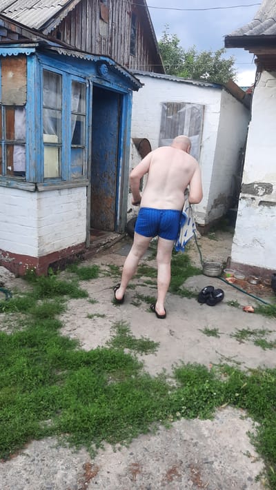 This media file captures a full-body shot of a bald man from behind, wearing blue checkered boxer briefs. He is standing on a concrete or paved surface in an outdoor setting, with a small amount of green grass growing through cracks in the ground. To the man's right, a pair of black flip-flops are visible on the ground. Behind him, to the left, is a weathered building with peeling blue paint on its facade, including a door and windows. The building appears to be somewhat dilapidated. The overall scene suggests a rustic or rural environment, possibly a backyard or side yard of a home. The lighting suggests it is daytime, and the cloudy sky in the upper left corner indicates overcast weather. There is no visible text in the image.