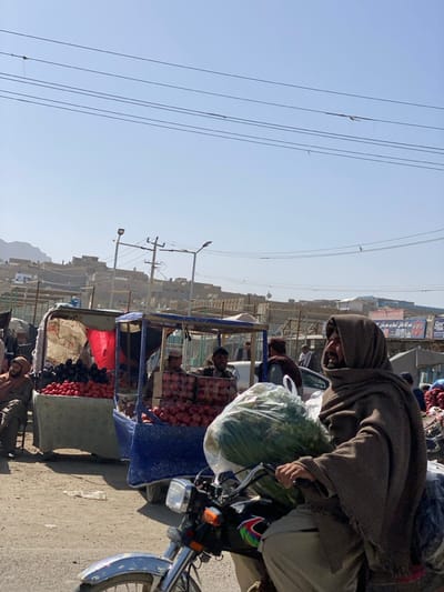 A man in a hooded garment rides a motorcycle, a large bag of greenery strapped to the front. He's positioned alongside a bustling outdoor market. Vendors with carts display produce, including apples, tomatoes, and more. The background shows buildings clinging to a hillside under a pale blue sky. Power lines crisscross the sky, adding to the urban landscape. The scene captures a typical day in a vibrant marketplace.