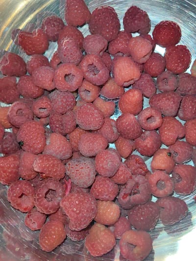 The image displays a close-up, overhead view of a metal bowl filled with fresh raspberries. The raspberries are various shades of pink and red, with some appearing slightly more translucent than others, indicating a range of ripeness. The surface of the berries is textured with small drupelets, characteristic of raspberries. The metal bowl is circular and has a brushed or ribbed surface, creating reflections and highlights that interact with the raspberries. There are no people, buildings, or specific landmarks visible in the image. The scene is a still life of fruit within a container, suggesting it could be in a kitchen or a place where food is prepared or stored. There is no information available to determine its location in Slavyansk-na-Kubani, Russia.