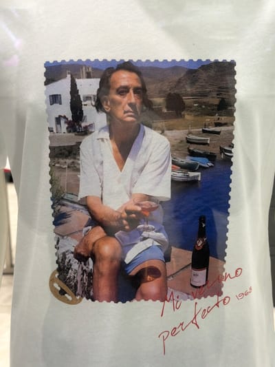 The image shows a postcard-style print of a man sitting by the water. He is fair-skinned with dark hair and is wearing a white shirt and blue swimming trunks. He is holding a glass in his left hand. Beside him is a bottle of what appears to be rosé wine. The background includes a body of water, boats, and buildings. Written in red ink on the white border of the print are the words "perfetto" and "Mio Veneto 1968". A perforated edge surrounds the print, giving it the appearance of a stamp or postcard. The image is presented on a white surface. There is no indication this is set in Novoaleksandrovka, Russia.
