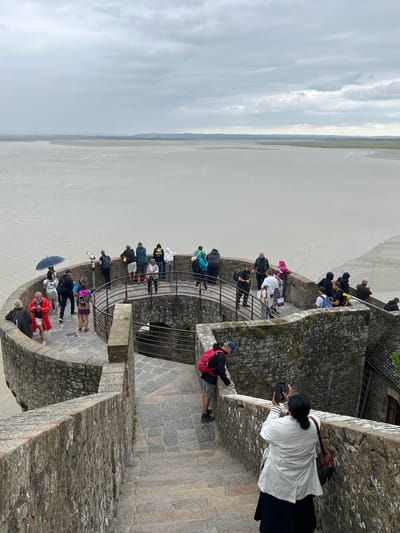 The image shows a group of tourists on a circular stone pathway overlooking a wide, muddy body of water under a cloudy sky. The pathway has a metal railing and is constructed with rough-hewn stones. Several people are standing and looking out at the water, while others are walking or taking pictures. Some individuals are holding umbrellas. A coin-operated telescope is visible on the pathway. The architecture suggests a historic site, consistent with the appearance of Le Mont-Saint-Michel, a tidal island and commune in Normandy, France, known for its medieval abbey. The overall mood is overcast and somewhat damp, as indicated by the presence of umbrellas and the muted colors of the landscape.