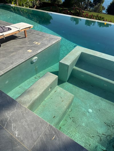The image shows a view of a swimming pool, likely in Vouliagmeni Municipal Unit, Greece, given the context of the surrounding landscape and water. The pool features an infinity edge, with clear turquoise water. Built-in steps with a textured finish lead down into the pool. The poolside is paved with large, dark gray stone tiles. On one side, there is a built-in seating area or shallow section of the pool. A wooden lounger with a white cushion and a dark gray towel is placed on the tiled deck. The water contains some debris, appearing to be leaves or plant matter. In the background, lush green foliage and trees are visible, with a glimpse of the sea and a bright blue sky. Sunlight casts shadows across the pool area.