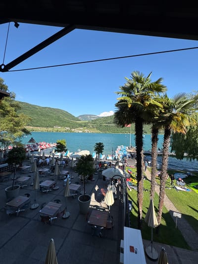 The image presents a vibrant, sun-drenched outdoor scene overlooking a lake in Kaltern an der Weinstraße, Italy. The perspective is from an elevated vantage point, looking across an outdoor dining area towards a large body of water and distant mountains.

In the foreground, a spacious paved patio hosts an array of outdoor dining tables and chairs, many shaded by light-colored parasols. Several patrons are seated, enjoying the weather. Large planters containing small trees are placed strategically around the seating arrangements. To the far left, a dark structural element of a building partially frames the shot.

The midground features the expansive, clear turquoise water of the lake. Along its shore, a lively recreational area is visible. A series of red and white paddle boats are docked, and people can be seen on the water, some on paddle boats, while others are engaging in water sports like windsurfing, with colorful sails and boards resting on a strip of green grass by the water's edge. Three tall palm trees with dense green fronds stand prominently, dividing the dining area from the lake and casting long shadows.

In the background, rolling green mountains rise from the opposite shore of the lake under a pristine, clear blue sky. The mountainsides show signs of cultivation or scattered buildings.

The scene depicts a leisurely day, likely midday or early afternoon, given the bright, direct sunlight and distinct shadows. The weather is clear and warm, contributing to a relaxed and enjoyable atmosphere. No specific text is clearly legible on any signs or objects within the image, though some tables have faint patterns or logos.