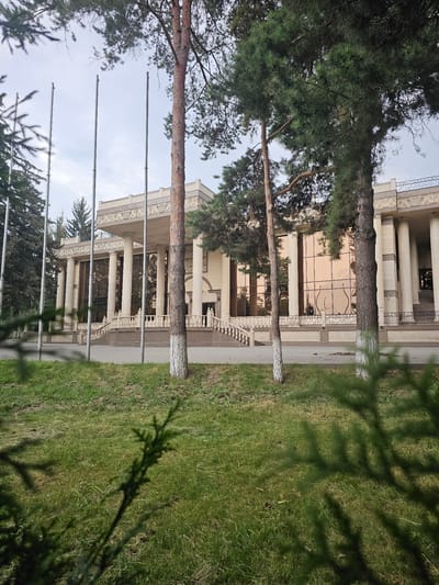 This is a vertical, full-shot photograph of a modern building with a grand entrance. The building features large, reflective windows that mirror the sky and surrounding trees. The facade is adorned with classical columns and decorative friezes. A manicured lawn with sparse trees surrounds the building, and a few flagpoles are visible in the foreground. The image appears to have been taken during the daytime, under a clear sky. The lighting suggests it's a bright day, possibly late morning or early afternoon. The overall scene conveys a sense of grandeur and serenity. There is no visible text in the image. The scene is likely a public building, such as a government office, museum, or convention center, in Chong-Aryk, Kyrgyzstan. The presence of greenery and the clear sky suggest pleasant weather.
