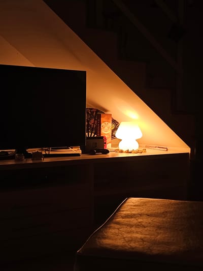 This media file presents an indoor, dimly lit scene characterized by warm artificial light. The central feature is a bright, white lamp, shaped like two connected ovals or a stylized 'M', which casts a strong, warm glow onto the surrounding light-colored walls.

Below the lamp, a box bearing the text "CHIVAS 12" is visible, alongside what appears to be a Nintendo Switch console, identifiable by its red and blue Joy-Cons. Behind these items, there is a patterned fabric or decorative paper. To the right of the lamp, a series of small, light-colored objects, possibly figurines or ornaments, are arranged vertically against the wall.

In the lower left foreground, a large, dark, rectangular object, likely a television screen or piece of furniture, partially obscures the view. On the right side of the image, a textured, light brown vertical panel or curtain is illuminated by the lamp's spill light, revealing its coarse surface. Various wires and electrical components are visible along the right edge, behind the large dark object.

The setting appears to be a nook or corner, possibly beneath a staircase or sloped ceiling, hinted at by the diagonal lines in the dark upper left portion. The scene is static, devoid of human presence or activity, and implies an intimate or cozy atmosphere, characteristic of nighttime or low-light conditions indoors. The visible text is "CHIVAS 12". The location context is Cabo Frio, Brazil.