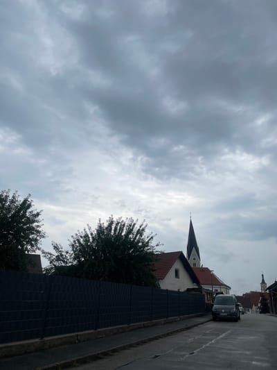 The image captures a view of a street in Geiselhöring, Germany, on a cloudy day. In the foreground, a dark fence lines the side of the road, which appears wet, suggesting recent rain. Several houses with red tiled roofs are visible behind the fence, with some trees also present. A prominent church steeple with a clock is visible in the middle ground. A dark gray van is parked on the right side of the street, facing forward. In the background, more buildings and another, smaller church steeple can be seen. The sky is heavily overcast with dark gray clouds.