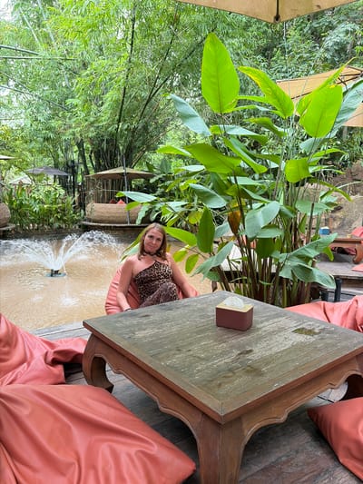 A woman is seated in a beanbag chair next to a wooden table and a fountain in Tambon Mae Raem, Thailand. She is wearing a leopard print one-shoulder jumpsuit and a choker necklace. The fountain is in a shallow pond surrounded by lush greenery and bamboo. The table is dark wood with ornate legs and has a tissue box on it. The beanbag chairs are a vibrant coral color and are scattered around the table. In the background, there are thatched umbrellas and seating areas, indicating an outdoor dining or lounge space. The overall setting appears to be a tropical resort or restaurant.