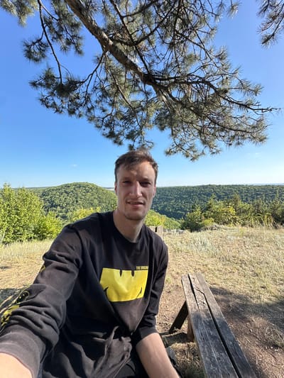 A young man with short brown hair and blue eyes is taking a selfie outdoors. He is wearing a black long-sleeved shirt with a yellow smiley face graphic and the text "THE ORIGIN" on it. He is sitting on a wooden bench with a weathered appearance. The background shows a lush green valley with rolling hills covered in trees, under a clear blue sky. Pine branches hang from above, framing the upper part of the image. The setting appears to be a scenic overlook, likely in or near the city of Nihyn, Ukraine, given the context of the prompt. The overall atmosphere is bright and natural, suggesting a daytime outdoor activity.