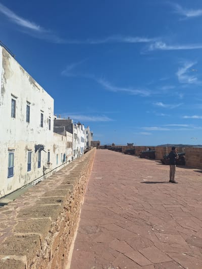 A person stands on a red-tiled promenade along a fortified wall in Essaouira, Morocco. The wall, made of rough-hewn brown stone, separates the promenade from a row of white buildings. The buildings, some appearing weathered and aged, have numerous windows with blue shutters. A sign for "RESTAURANT La Lune" is visible on one of the buildings. Along the fortified wall, several cannons are positioned, facing outwards towards the sea. The sky above is a vibrant blue with scattered wispy clouds, and several seagulls are in flight. The promenade is paved with irregular, reddish-brown tiles, and a shadow of the standing person is cast upon it. The overall scene suggests a historical coastal fortification with a mix of architecture and a clear, sunny day.