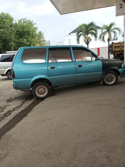 A turquoise and dark gray minivan, the "Kijang" model, is parked on a concrete surface at a gas station in Tocina, Indonesia. The minivan is positioned in profile, facing right, with its rear passenger door slightly ajar. The vehicle exhibits some wear and tear, with dirt visible on its windows and a mismatch in paint color on the front fender. The minivan's wheels are a notable feature, with intricate gold-colored rims.

To the left of the minivan, another vehicle, possibly a white SUV, is partially visible, also parked. Behind the vehicles, lush green trees and palm trees frame the scene. In the background, a white building with a dark roof stands out, with a yellow truck visible to its right. A portion of the gas station's canopy, with a red number "2" displayed on a white sign, overhangs the scene from the top. The concrete ground shows signs of moisture, with dark streaks possibly indicating oil or water spills. The sky above is overcast.