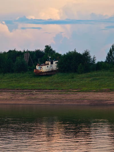 The image depicts a wide shot of a riverbank with a grounded boat. The boat, an old and rusty white vessel with some orange rust visible, is partially embedded in the grassy slope. It is surrounded by lush green trees and vegetation. A wooden structure, possibly a loading ramp or scaffolding, is visible to the left of the boat, leaning against the trees. The foreground is dominated by the calm, reflective surface of a river, with the sandy bank of the opposite shore visible in the middle ground. The sky above is filled with soft, diffused clouds, suggesting it is either early morning or late evening, with a hint of warm light in the upper right corner. The overall mood is peaceful and somewhat melancholic, with the abandoned boat being the central focus of attention. There is no visible activity or people in the scene. The visible text on the boat is "CK".