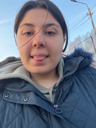 The image displays a close-up, chest-up outdoor portrait of a young adult female in Talin, Armenia. She has dark hair, a medium complexion, and dark eyes. A single strand of dark hair crosses her face over her right eye and nose, and a small, dark mole is visible above her right eyebrow. Her mouth is open, and her tongue is extended forward. She is dressed in a light gray hooded sweatshirt, layered under a dark blue quilted winter jacket. The jacket features a visible metallic zipper, partially unzipped, and metallic snap fasteners on the front placket. A dark blue faux fur trim is discernible around the jacket's hood/collar area on the subject's right side. The background consists of a clear, light blue sky, indicating daylight. To the upper right, a utility pole supports multiple utility lines and an attached street light fixture. In the distant background, a line of deciduous trees or shrubs, devoid of leaves, suggests a colder season.