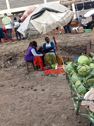 In Keekonyokie ward, Kenya, an outdoor market scene unfolds on unpaved, uneven ground. In the mid-ground, two adult women and a child are positioned near a display of produce. The woman on the left, wearing a purple vest, white top, and red skirt, sits on a wooden stool, with her left arm partially around the child. The child, in a dark top, is seated between the two women. The woman on the right, in a blue top and green patterned skirt, is seated lower, with arms crossed, facing the other woman. Piles of red tomatoes are arranged on a blue fabric on the ground in front of them, with some in a purple plastic bowl and a green basket, beside which a price sign is visible. A yellow plastic container and a stick with a red attachment are nearby. The ground in the immediate foreground is bare earth, littered with several small, dark, elongated objects resembling animal droppings. To the right, a green metal handcart is laden with numerous heads of green cabbage, with a cardboard sign attached to its frame. In the background, several individuals move amidst other makeshift market stalls, including a large, off-white tarp structure supported by poles, and a partial view of a concrete block building wall. The overall environment suggests an active, open-air commercial area.