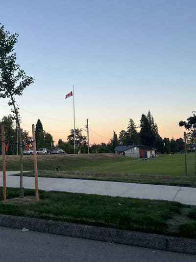 This media file depicts an outdoor scene at dusk in North Vancouver, Canada. The sky transitions from a pale blue at the top to hues of orange and pink near the horizon, indicating the sunset. In the distance, a Canadian flag flies at half-mast on a tall flagpole, a solemn detail that suggests a moment of remembrance or mourning.
The scene includes a well-maintained green lawn adjacent to a paved walkway. A small building with a dark roof and light-colored walls is visible in the mid-ground, possibly a community center or park facility. Several cars are parked in a line near the building, suggesting some activity or event is taking place. Trees, some with dark foliage, line the background, adding depth to the scene. In the foreground, a few trees with lighter green leaves are partially visible, as well as some wooden posts with orange netting, which could be part of a temporary barrier or construction.
The overall mood is quiet and peaceful, amplified by the soft lighting of dusk. The presence of the flag at half-mast adds a touch of somberness to the otherwise serene setting. There is no visible text in the image.