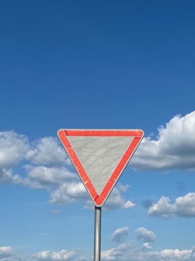 The image depicts a triangular traffic sign with a red border against a bright blue sky filled with fluffy white clouds. The sign is mounted on a metal pole, which extends horizontally across the lower right portion of the frame. The surface of the sign is a weathered grey and shows signs of cracking and wear along the red border. The sky is clear and vast, with sunlight illuminating the clouds and the sign. The orientation of the image is vertical. There is no visible text on the sign itself. The setting appears to be an outdoor location, likely a roadside in Belarus, given the provided location context. The overall mood is serene and expansive, with the vastness of the sky dominating the scene.