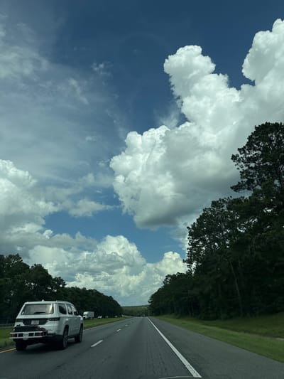 The image captures a scene from a vehicle traveling on a highway, possibly in the vicinity of Midway, United States, given the context of a road and trees. The perspective is from inside a moving car, looking forward and slightly to the left. The sky is a dominant feature, with a vibrant blue expanse interspersed with large, fluffy white cumulus clouds. These clouds are dramatic and well-defined, suggesting a partly cloudy day with potential for fair-weather cumulus formation.

In the foreground, on the left side of the highway, a white SUV is visible. It appears to be a newer model, possibly a Grand Wagoneer, as indicated by lettering on the rear. The SUV has a cargo carrier attached to its hitch, suggesting it is equipped for travel or hauling. The vehicle is in motion, contributing to the sense of a journey.

The highway itself is paved, with clear white lane markings indicating multiple lanes of travel. The road stretches into the distance, flanked by dense green trees on both sides. These trees, primarily evergreens with some deciduous varieties, create a natural barrier along the highway, suggesting a rural or suburban setting rather than a dense urban environment. The trees on the right appear taller and more prominent, with some of their branches reaching towards the sky.

Further down the highway, other vehicles are visible as smaller shapes, indicating ongoing traffic. The overall impression is one of a road trip on a clear, bright day with dynamic cloud formations overhead. The environment appears to be a typical American highway landscape, likely in a region with significant tree cover.