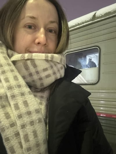 A person with light brown hair and a patterned scarf is facing the camera. A dark-colored jacket is draped to the right. Behind them is the side of a white, possibly metallic structure with narrow vertical ridges. A small window is visible, showing a dark figure inside the structure, giving the impression they might be on a vehicle or within a building. The sky in the upper left corner has a pale, lavender hue.