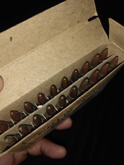 The image depicts an open, light brown cardboard ammunition box held by an individual's fingers, visible at the bottom-left. The box contains three visible rows of metallic cartridges, each with a brass-colored casing and a conical, copper-jacketed bullet. White, powdery residue or debris is present on the cartridges and within the cardboard dividers separating them. Some cartridges display discoloration or oxidation. The cardboard box exhibits minor surface wear. The background is dark and featureless. The specific location within the city of Tas-Kop', Kazakhstan, is not discernible from the image.