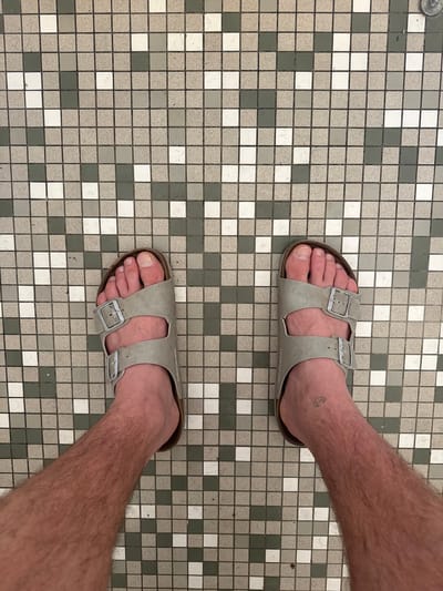 A top-down, medium shot shows two feet wearing grey Birkenstock sandals on a mosaic tile floor. The tiles are square and arranged in a grid pattern, with a mix of white, light gray, and dark gray tiles. Some of the tiles appear to be speckled. The feet are positioned diagonally from the top right to the bottom left of the frame. The left foot, closer to the top of the image, has a small, faint tattoo of a crescent moon on the ankle. The right foot is closer to the bottom of the image. The background is exclusively the tiled floor. There is no discernible time of day, weather, or specific emotion conveyed. The location is identified as Bellelay, Switzerland. The text "BIRKENSTOCK" is faintly visible on the sole of the sandal on the right foot.