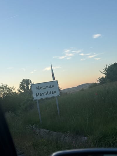 The image shows a road sign indicating the entrance to the village of Meshtitsa in Bulgaria. The sign features the village name in both Cyrillic (Мещица) and Latin script (Meshtitsa). A Bulgarian flag is visible near the sign. The background includes a grassy field, trees, a hill, and a sky with some clouds. The photo appears to have been taken from a vehicle, with part of the car visible in the edge of the image.