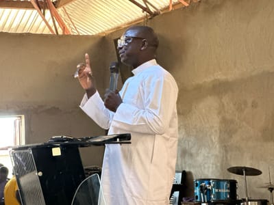 A Black man wearing glasses and a white clerical garment stands in what appears to be a church, holding a microphone and speaking with his index finger raised. To the left of the man, a black keyboard and what looks like a glass shield are visible, with two other people sitting in the background. To the right of the man, a blue drum set with the words "TBC M/KAJURU" printed on it, as well as a cymbal, are present. The room has rough, concrete walls and a ceiling with exposed wooden beams and corrugated metal roofing. The man is positioned in the center of the frame, facing towards the right. The lighting is dim, suggesting an indoor setting.