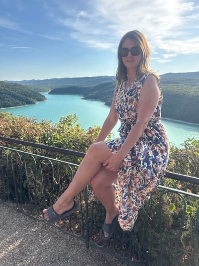 A woman is sitting on a black metal railing, her legs crossed and her body angled towards the right. She is wearing sunglasses, a floral print dress, and dark grey slides. She is positioned in front of a scenic overlook with a wide body of turquoise water, likely a reservoir, bordered by lush green hills and a bright blue sky with scattered clouds. The foreground consists of a gravel path and some vegetation. The location is identified as Moirans-en-Montagne, France, suggesting this is a viewpoint overlooking the Lac de Vouglans, a reservoir in the Jura region.