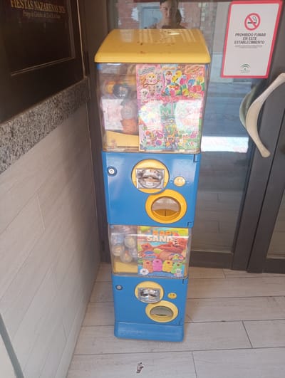 The image shows a vending machine for toys located in the city of Priego de Córdoba, Spain. The vending machine is blue and yellow, with three transparent compartments filled with small toys. The top compartment has a colorful sticker with cartoon characters and the text "SUPER MIX". The middle compartment has a coin slot and a dispensing chute. The bottom compartment contains more toys and a sticker with the text "KAPY SAND".
The vending machine is positioned next to a glass door, through which a woman can be seen in the background. To the left of the vending machine, there is a dark-colored door with a sign that reads "FIESTAS NAZARENAS 2026". To the right of the vending machine, there is a white sign with a red border that says "PROHIBIDO FUMAR EN ESTE ESTABLECIMIENTO". The floor is tiled with light-colored wood-patterned tiles.