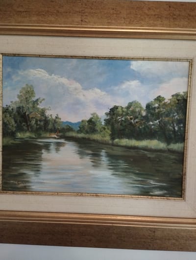 This is a framed oil painting depicting a serene river scene with lush green trees lining the banks. The sky is a bright blue with fluffy white clouds. In the distance, there appear to be rolling hills. The water reflects the sky and the surrounding foliage, creating a sense of depth and tranquility. There is a small red figure in a boat on the river, suggesting human presence and activity within the natural landscape. The painting is housed in a decorative frame, with a golden outer frame and a lighter inner mat. The style of the painting is impressionistic, with visible brushstrokes that contribute to the overall artistic quality of the scene. The image is not specific to Zikhron Yaakov, Israel; it is a general landscape painting.