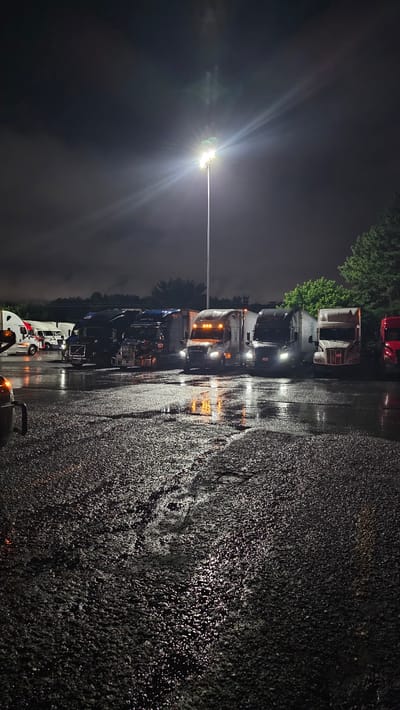 A long shot photograph captures a dimly lit parking lot at night, likely a truck stop or rest area, in Cartersville, United States. The ground, a mixture of asphalt and gravel, is wet and reflects the overhead lighting from a tall pole in the background, creating shimmering patterns. A line of semi-trucks is parked, their headlights and interior lights illuminating portions of the vehicles. The sky above is dark and cloudy, with the bright light from the pole piercing through. The atmosphere is quiet and still, suggesting a pause in the nocturnal journey of the trucks and their drivers.
