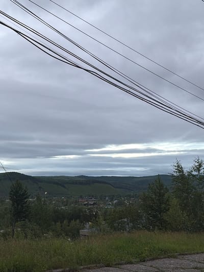 The image is a medium shot of a cloudy sky over a rural landscape near Skovorodino, Russia. In the foreground, power lines crisscross the frame against the sky. Below the sky, rolling hills covered in dense green vegetation are visible. A small village with scattered buildings and roads is nestled in the valley between the hills. The overall atmosphere is overcast and tranquil.