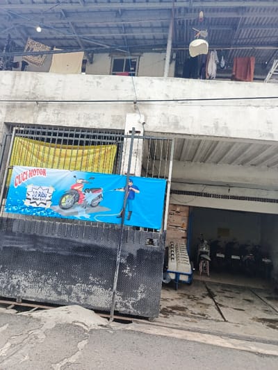 A two-story building with clothes hanging on the upper level. A blue banner advertising "CUCI MOTOR" (motorcycle wash) is prominently displayed in front of a gate. Several motorcycles are parked in an open garage area. The ground floor is covered in a rough cement. The building appears to be in a densely populated, possibly urban area.